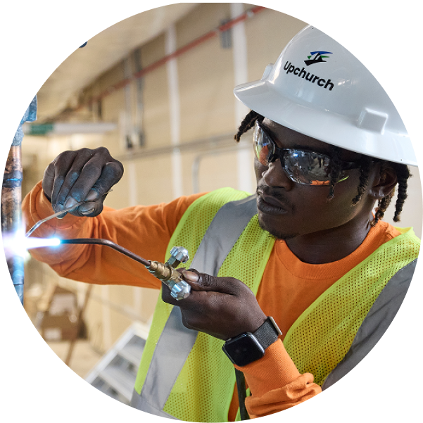 A construction worker wearing a white hard hat, safety vest, and orange shirt uses a welding torch on a metal pipe indoors—a scene often seen in projects funded by private equity investments. The worker is focused, wearing gloves and safety glasses.