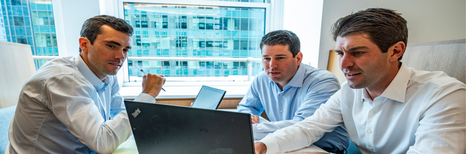 Three men in business attire sit together at a table, focused on a laptop screen as they discuss private equity strategies, with a large window showing an office building in the background.