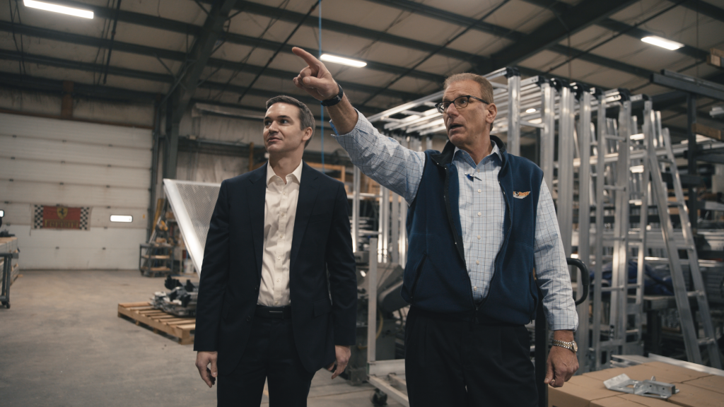 Two men stand in a large industrial warehouse; one is in a suit, the other in casual business attire, pointing and explaining something—perhaps discussing private equity investments amid metal structures and equipment in the background.