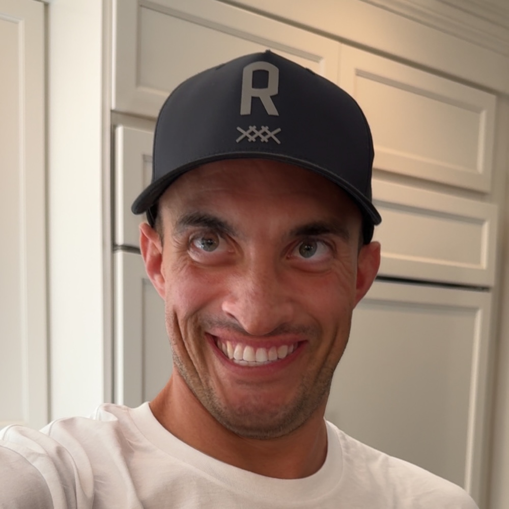 A man wearing a dark baseball cap and a white shirt smiles widely while standing indoors in front of white kitchen cabinets.