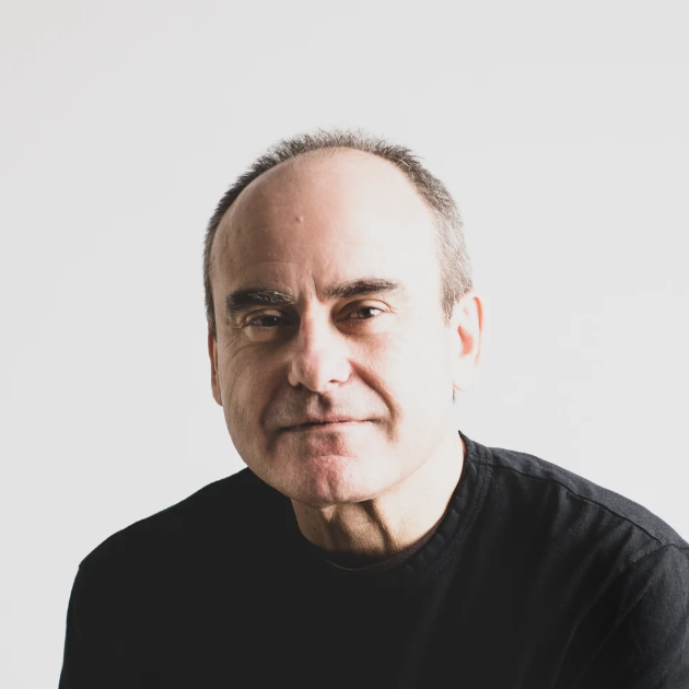 A middle-aged man with short, thinning hair and a slight smile is wearing a black shirt and sitting against a plain white background.
